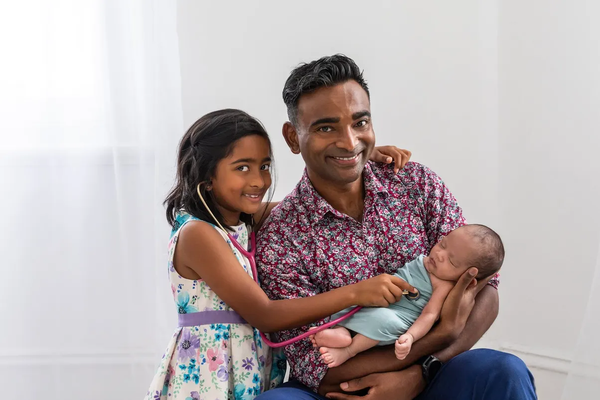 Dr Jubal John holding a newborn while his daughter listens with a toy stethoscope